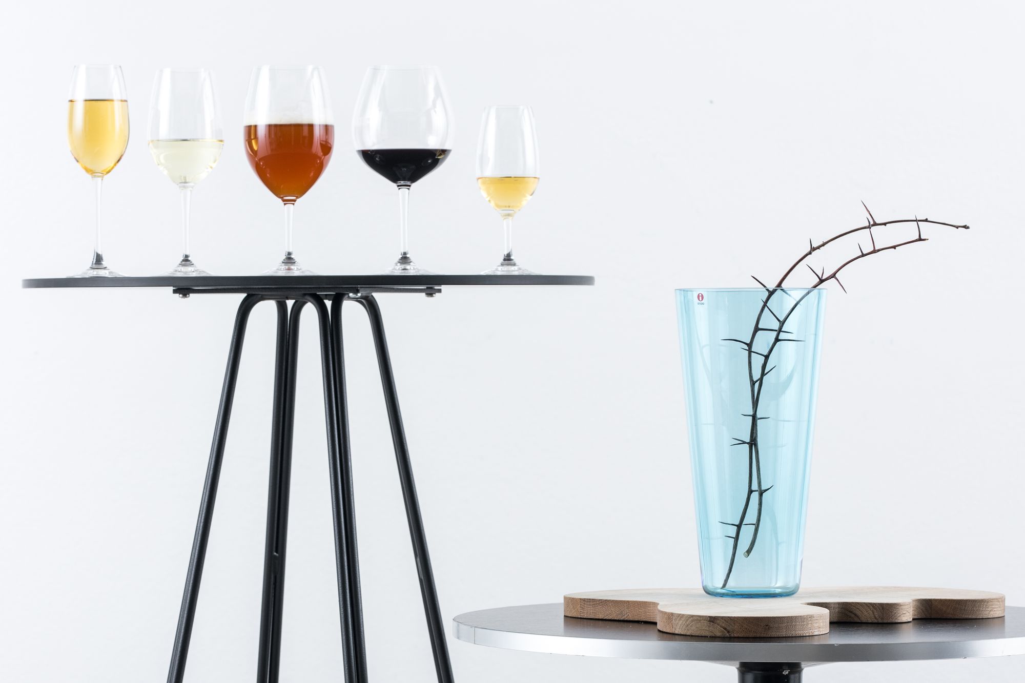 Viisi erilaista lasia juomineen korkealla tarjoilupöydällä. Pöydän vieressä moderni ja tyylikäs Iittalan maljakko, jossa kaksi oksaa. 