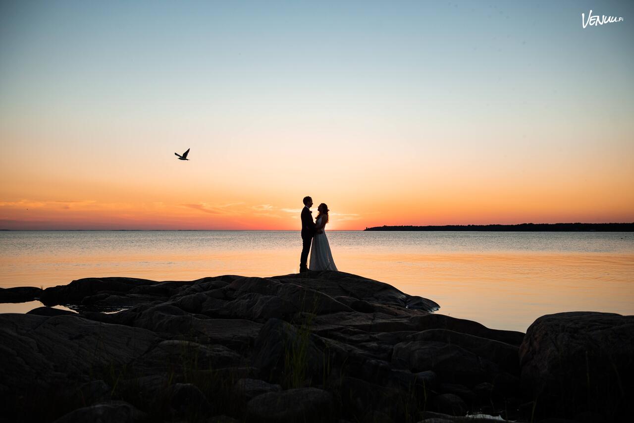 Aarographyn mieletön hääkuva parista kallioilla, joiden takana on upean punertava auringonlasku joka laskee meren taakse